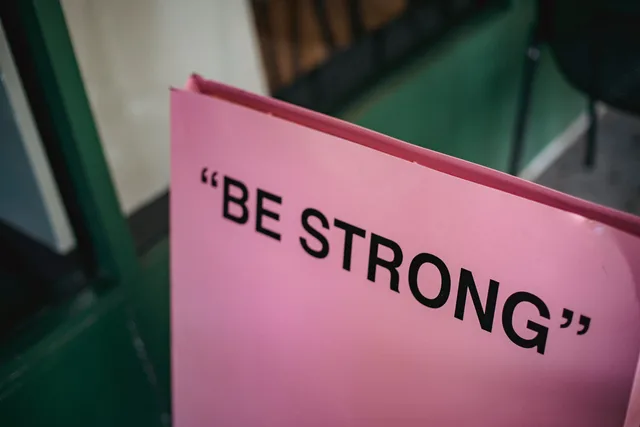 From above of pink signboard with black motivational phrase Be strong on street near green facade of building in city with blurred background. Werbemittel für kurze Aufmerksamkeitsspannen gestalten